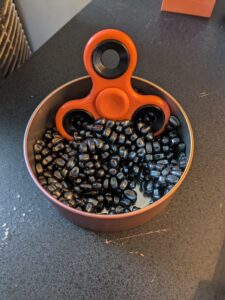 An orange fidget spinner in a container of small black fidget magnets. 